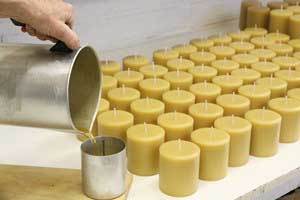 a candle maker pouring beeswax into a pillar candle mold with a table full of the finished candles in the background