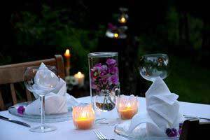 two burning pure beeswax candles in glass holders on a crisp white dining table set up outside with vibrant purple flowers